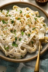 High protein creamy fettuccine Alfredo pasta topped with parsley and black pepper, served on a rustic plate with a golden fork.