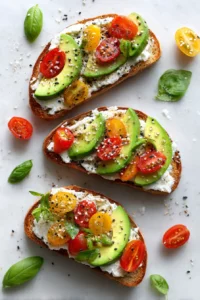 Overhead view of avocado toast topped with ricotta cheese, cherry tomatoes, and basil leaves