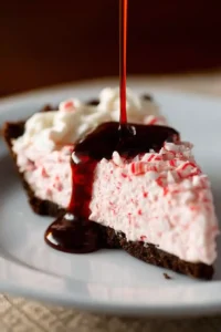 Slice of candy cane pie with a chocolate crust, whipped cream, crushed peppermint, and rich chocolate syrup being poured over the top.