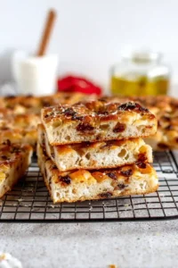 Golden brown cheesy focaccia bread topped with caramelized onions and herbs, cooling on a wire rack.